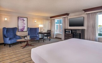 Habitación de hotel con una cama grande, dos sillones, un escritorio con silla, un televisor de pantalla plana y una ventana con cortinas.