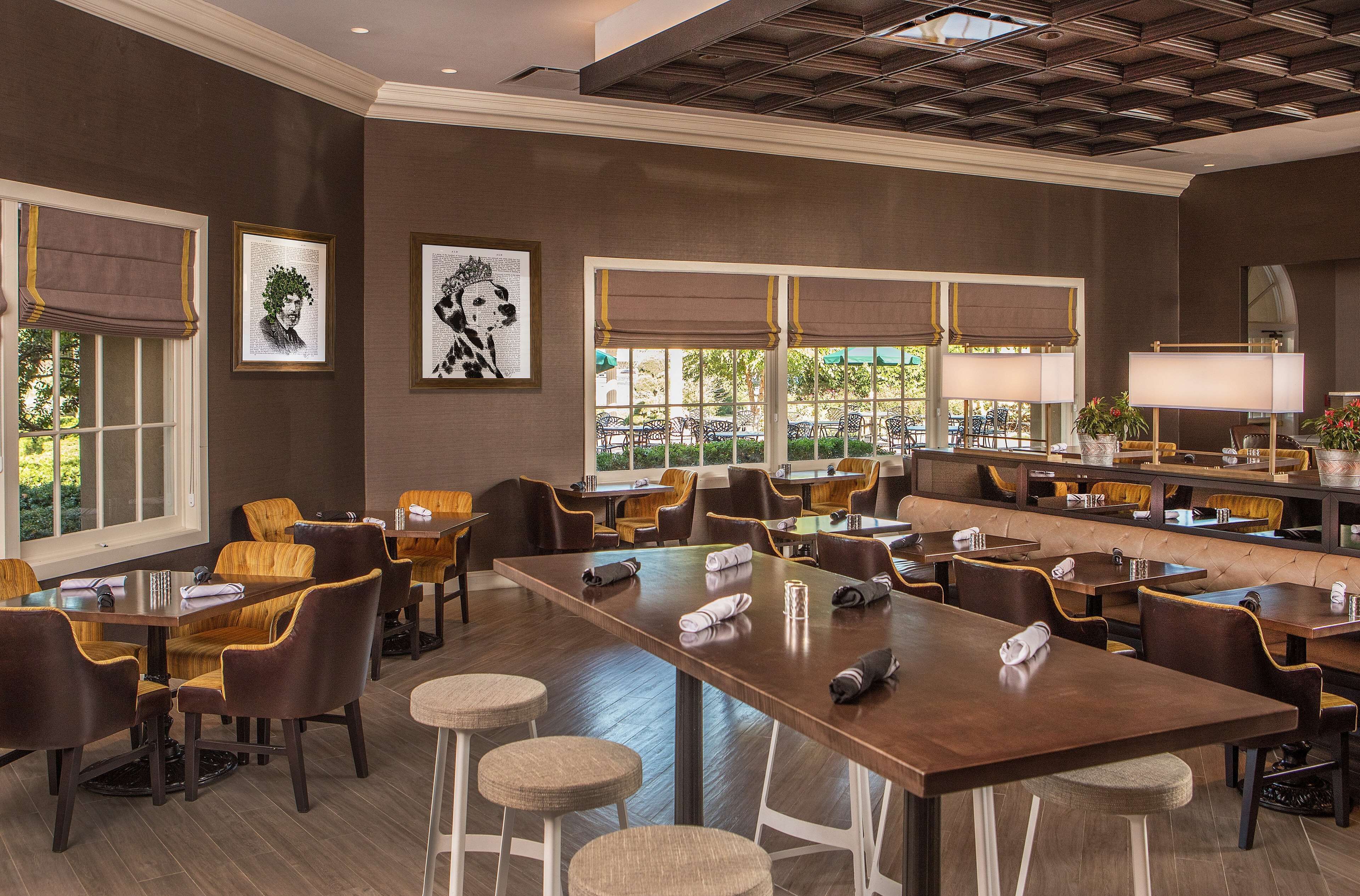 A spacious restaurant with high tables and comfortable seating, large windows with curtains, and framed artwork on the walls.
