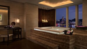 A modern bathroom with a large bathtub, wooden accents, a city view, and a vanity with a mirror and lamp. - The Peninsula Hong Kong (Kowloon)