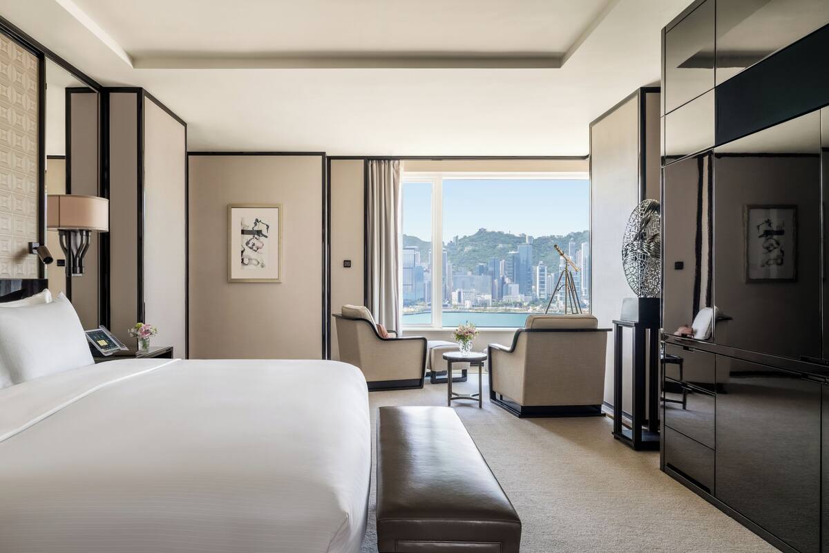 A hotel room with a large bed, a desk, a chair, a small table, and a view of a cityscape.