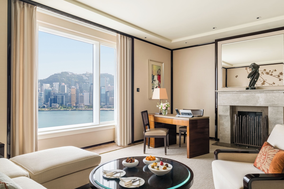 A hotel room with a desk, chairs, a dining table with food, a fireplace, and a large window with a city view.