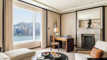 Quarto de hotel com escrivaninha, cadeiras, mesa de jantar com comida, lareira e uma grande janela com vista para a cidade.