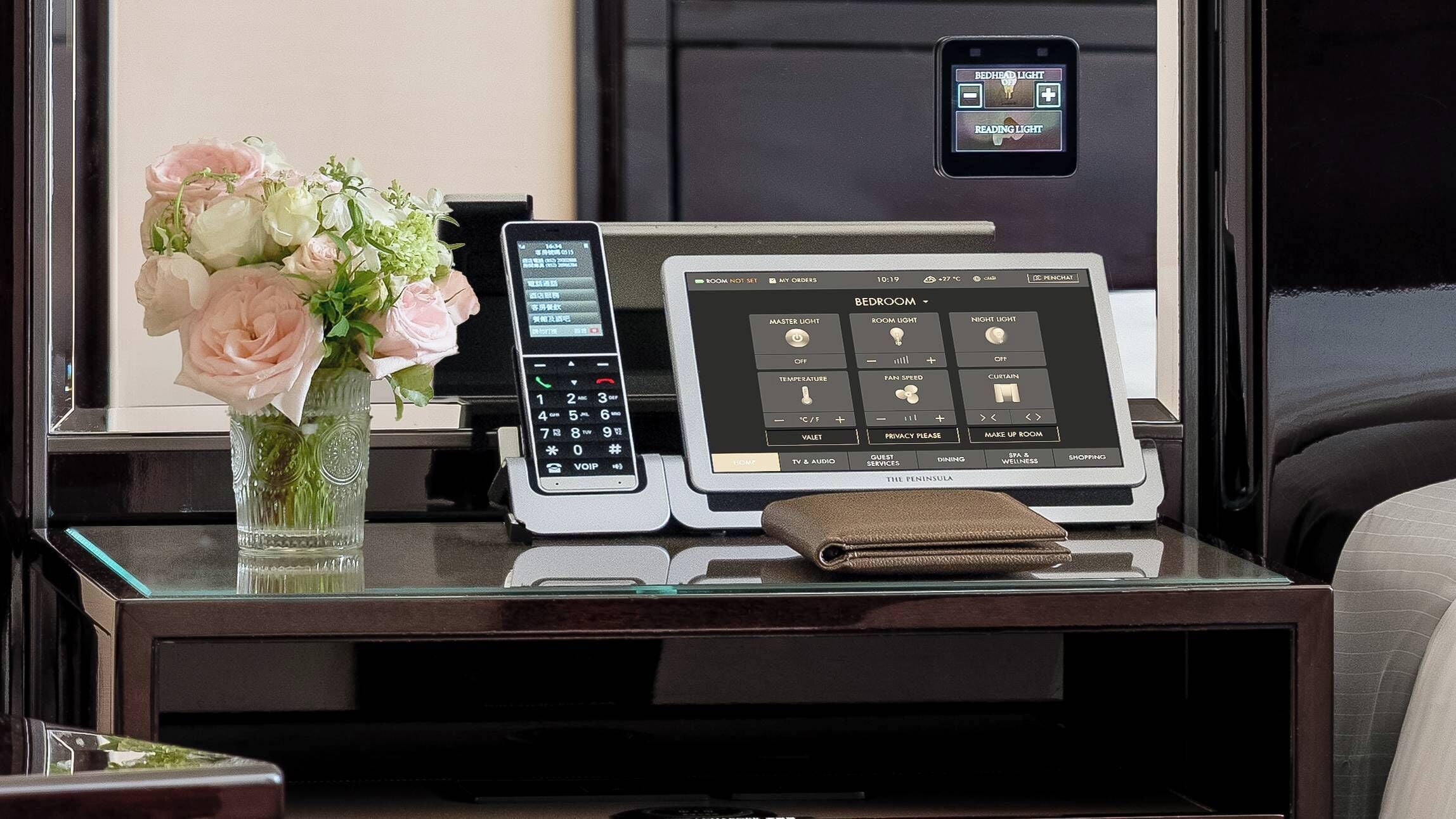 A hotel room control panel with a digital interface for room settings, a vase with flowers, and a small leather bag on the table.