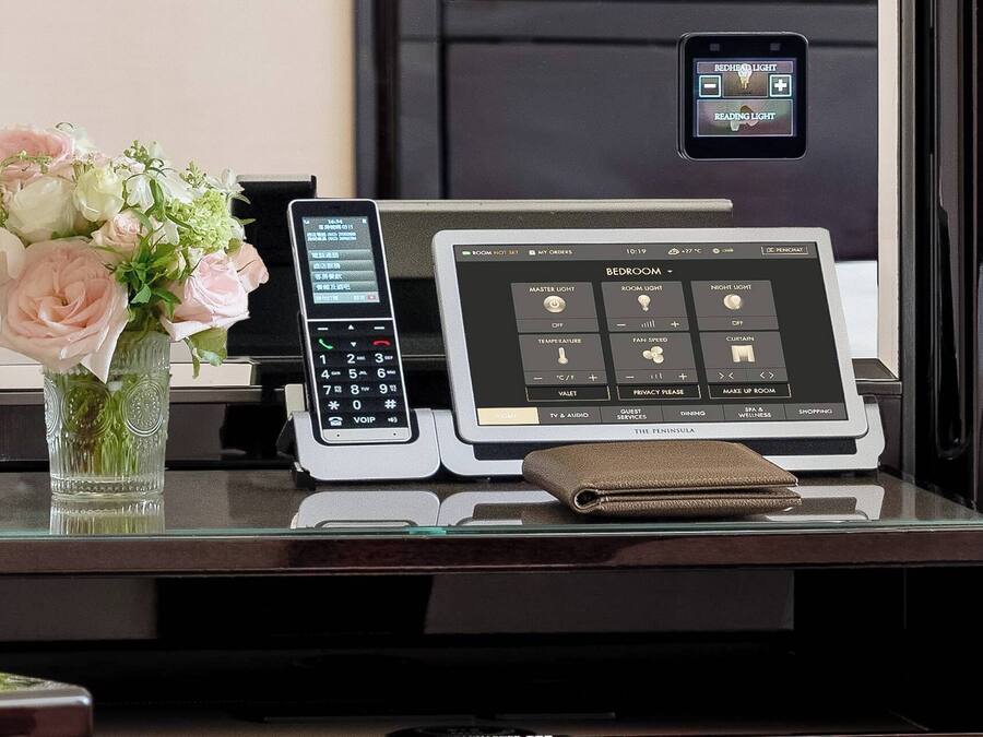 A hotel room control panel with a digital interface for room settings, a vase with flowers, and a small leather bag on the table.
