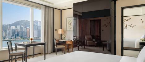 A hotel room with a large bed, a desk with a chair, a small table with a chair, and a view of a cityscape through large windows.