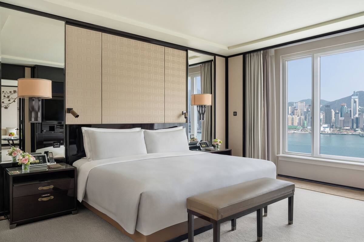 A hotel room with a large bed, a bench, a nightstand with a lamp, a TV, and a view of a cityscape.