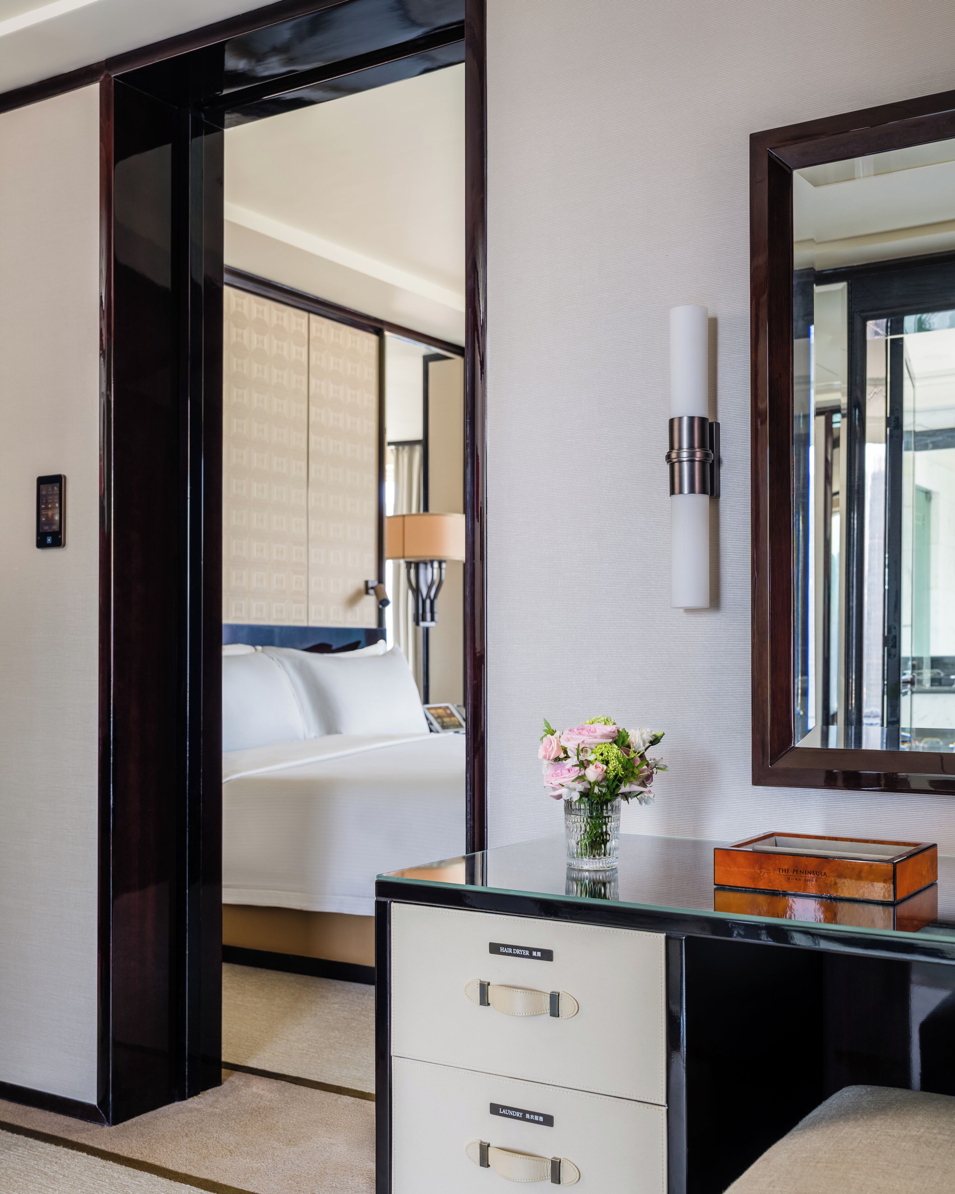 A hotel room with a bed, a desk with drawers, a mirror, and a vase with flowers.