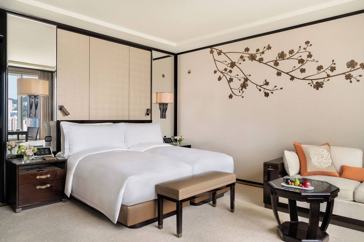 A hotel room with a large bed, a bedside table with a lamp, a small table with fruit, a sofa with cushions, and a decorative wall piece.