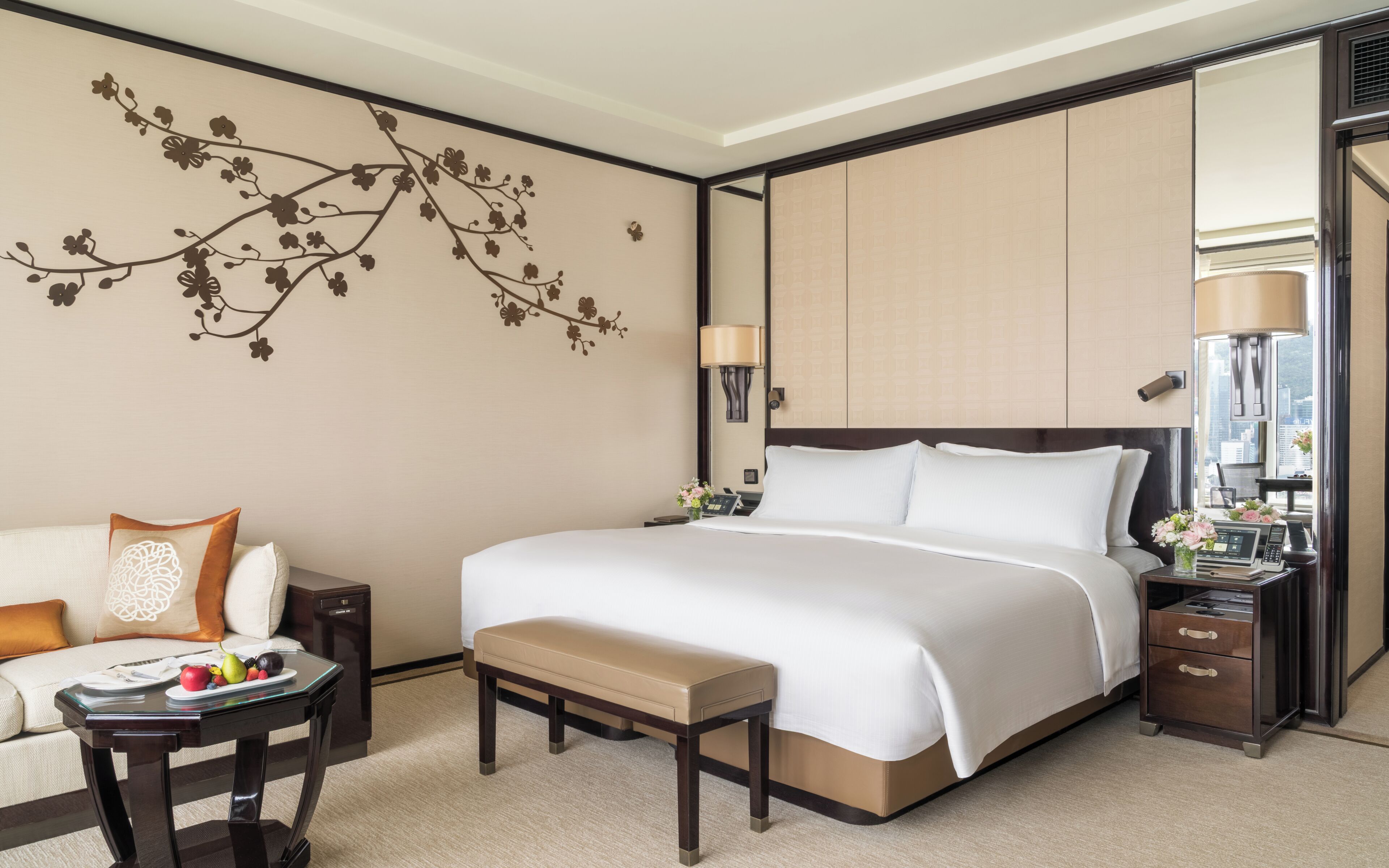 A hotel room with a large bed, a bedside table with a lamp, a small table with fruit, a bench, and a decorative wall piece.