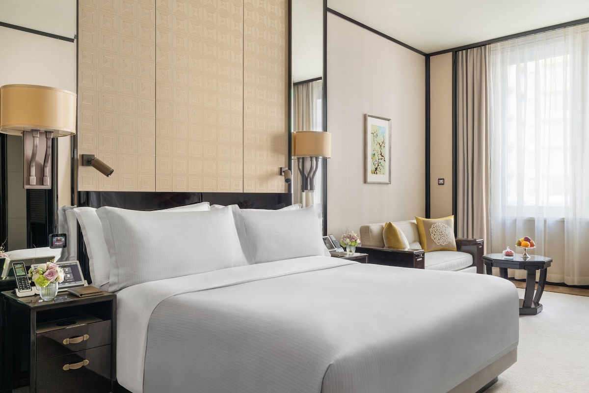 A hotel room with a large bed, bedside tables, a desk, a chair, a window with curtains, and a wall-mounted artwork.