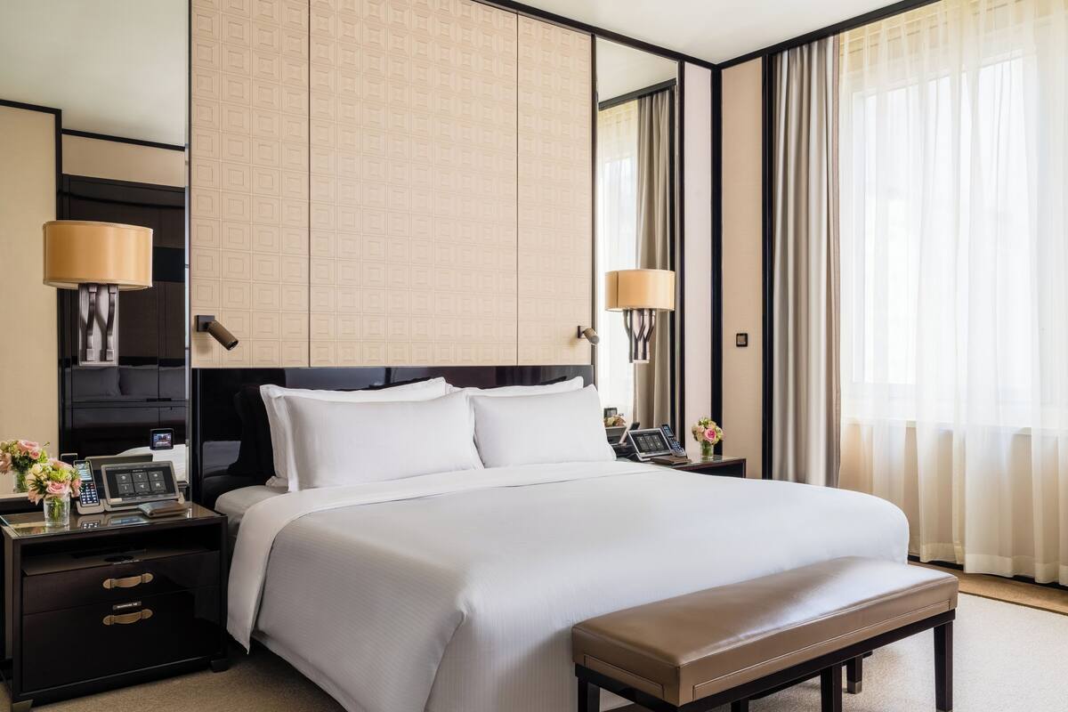 A hotel room with a large bed, bedside tables, a desk, a lamp, a mirror, and a window with curtains.