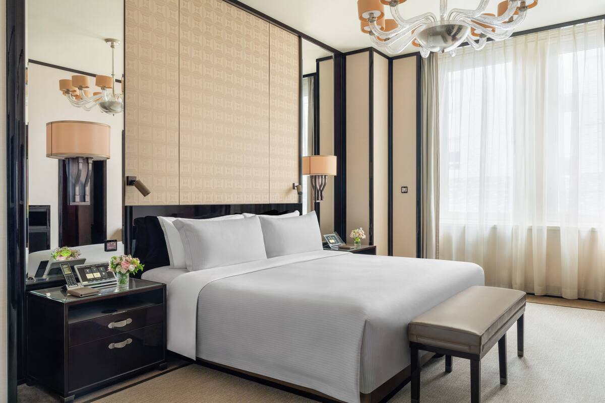 A hotel room with a large bed, bedside tables, a desk, a chair, a lamp, a chandelier, and a window with curtains.