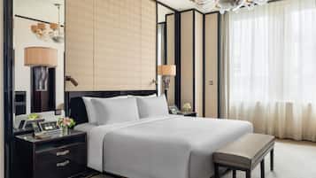 A hotel room with a large bed, bedside tables, a desk, a chair, a lamp, a chandelier, and a window with curtains.