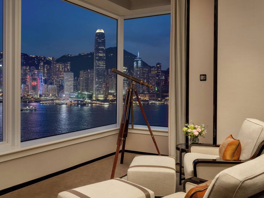 A hotel room with a large window overlooking a cityscape at night, featuring a telescope and seating area.