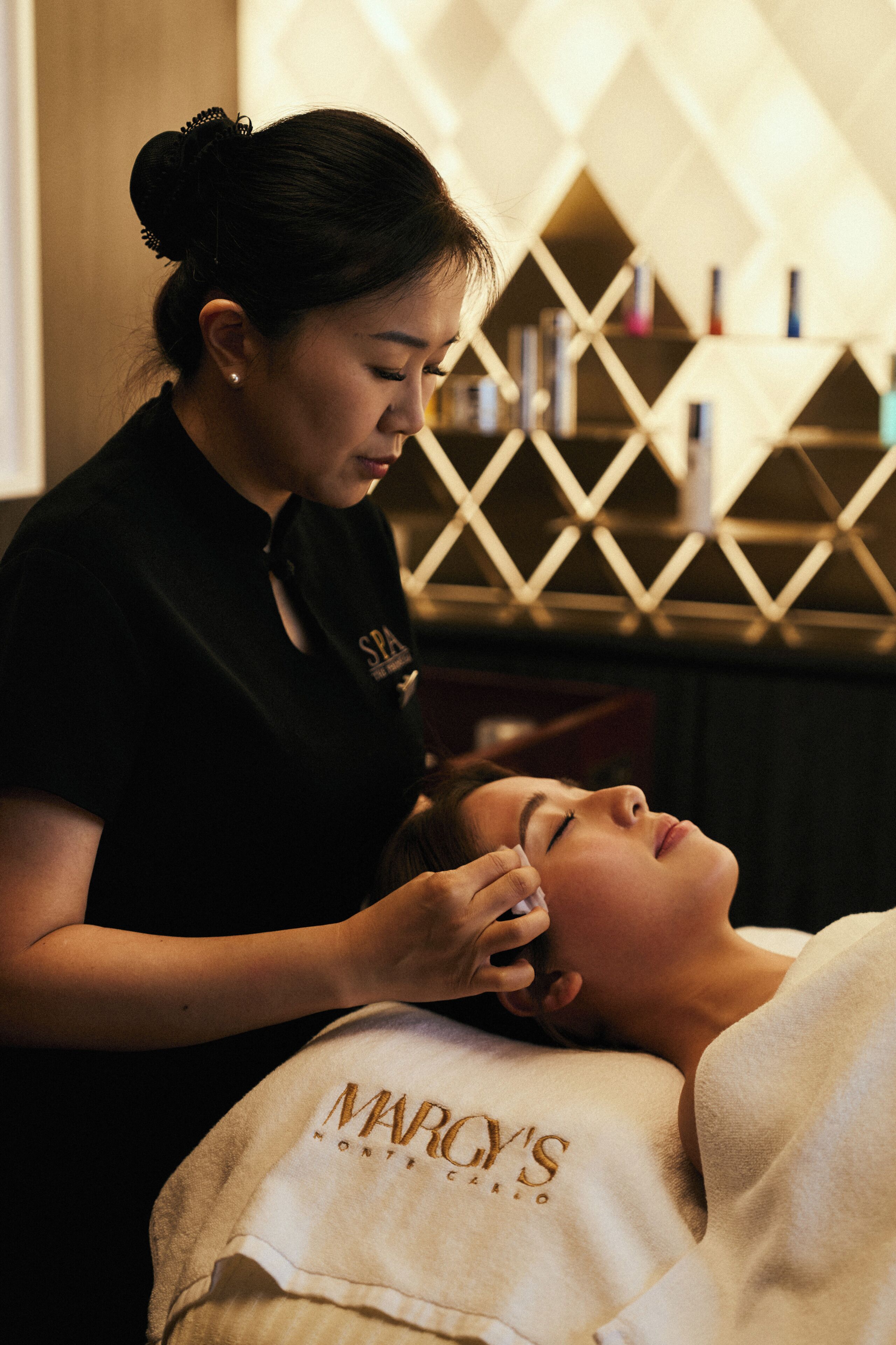 Uma pessoa recebendo tratamento facial em um spa, com parede e prateleiras decoradas com padrão geométrico ao fundo.
