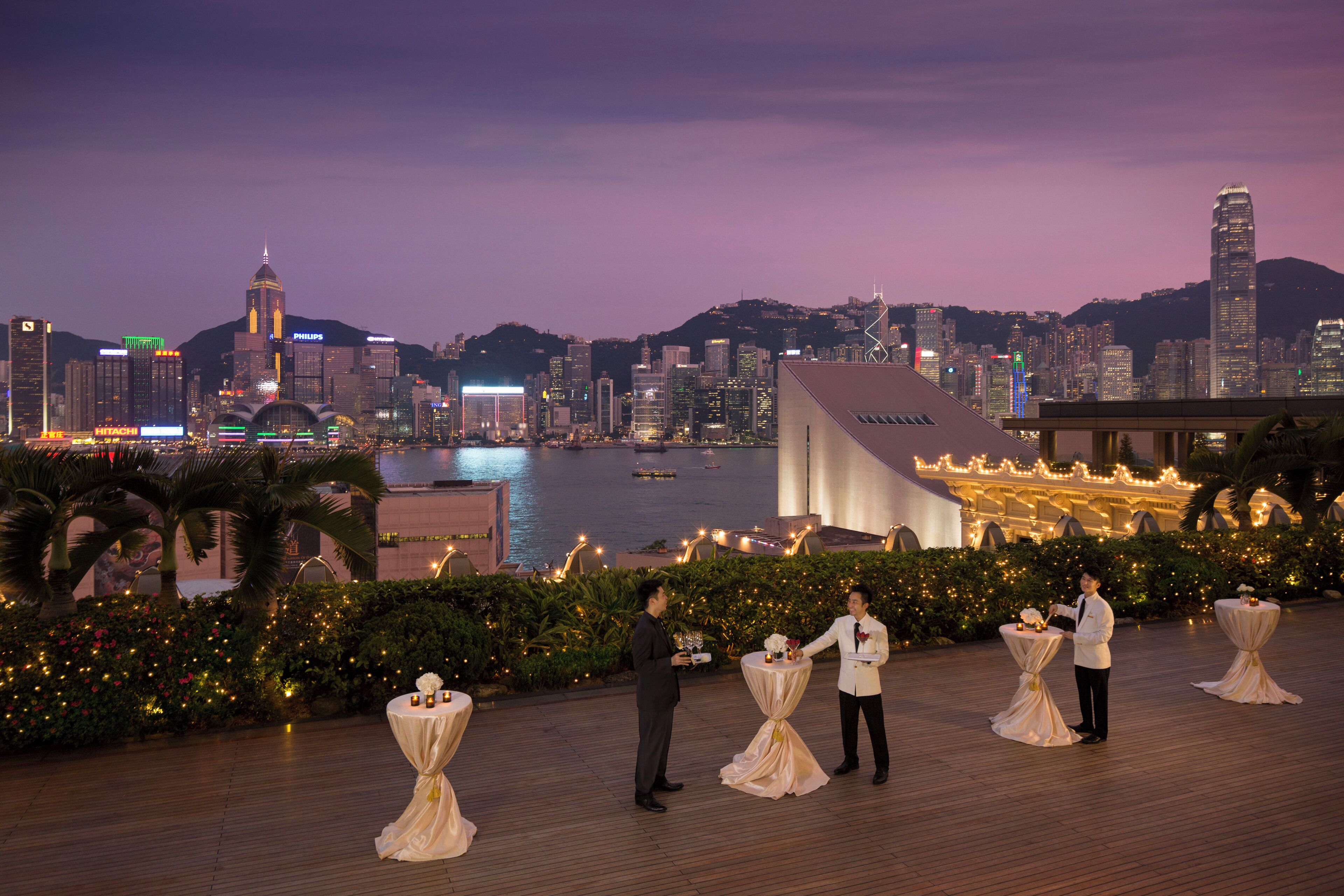 A waterfront venue with tables set for a reception, overlooking a city skyline at dusk.