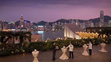 A waterfront venue with tables set for a reception, overlooking a city skyline at dusk.