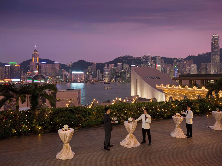 A waterfront venue with tables set for a reception, overlooking a city skyline at dusk.