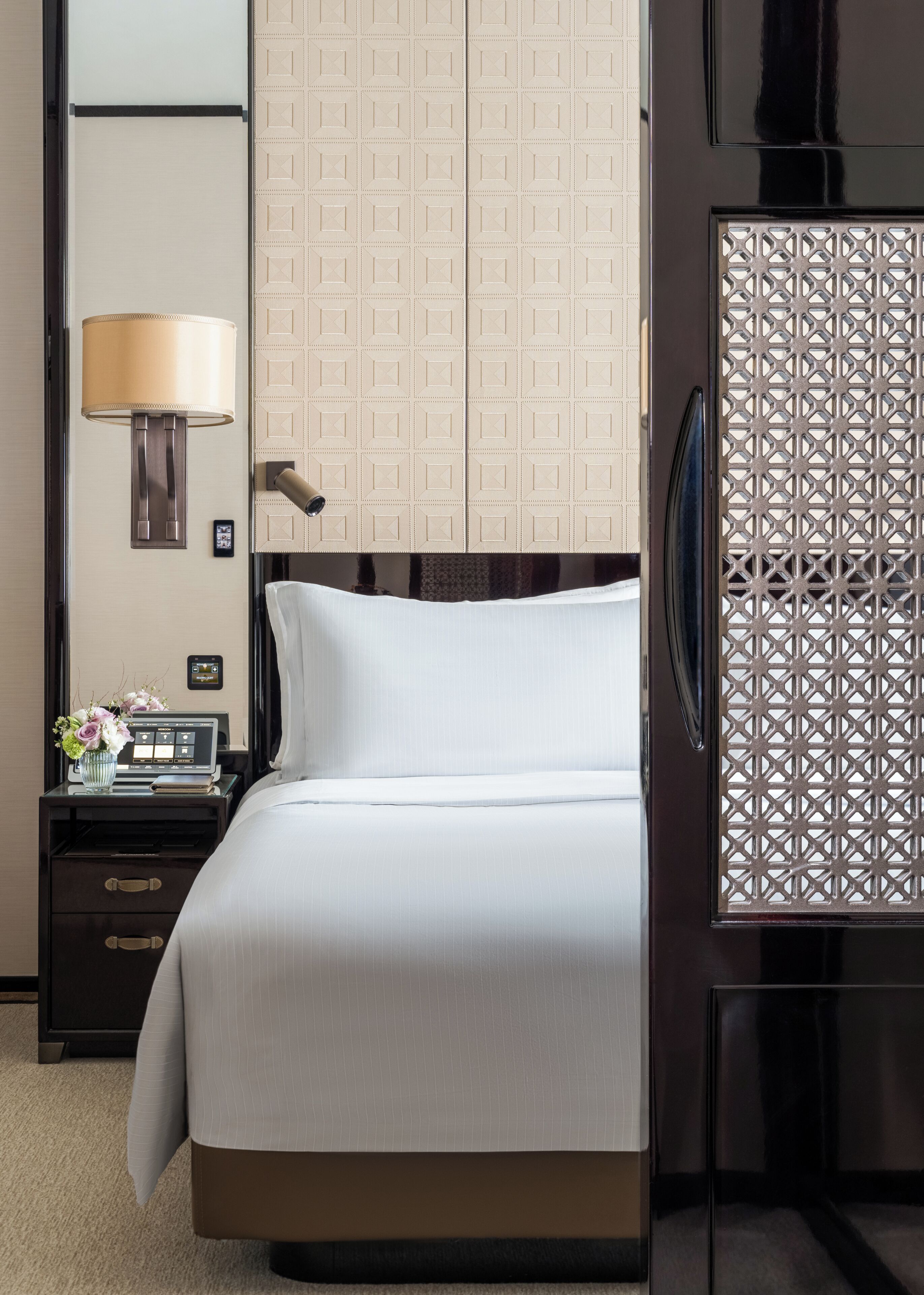 Quarto de hotel com uma cama, mesa de cabeceira com abajur, televisão e um biombô decorativo.
