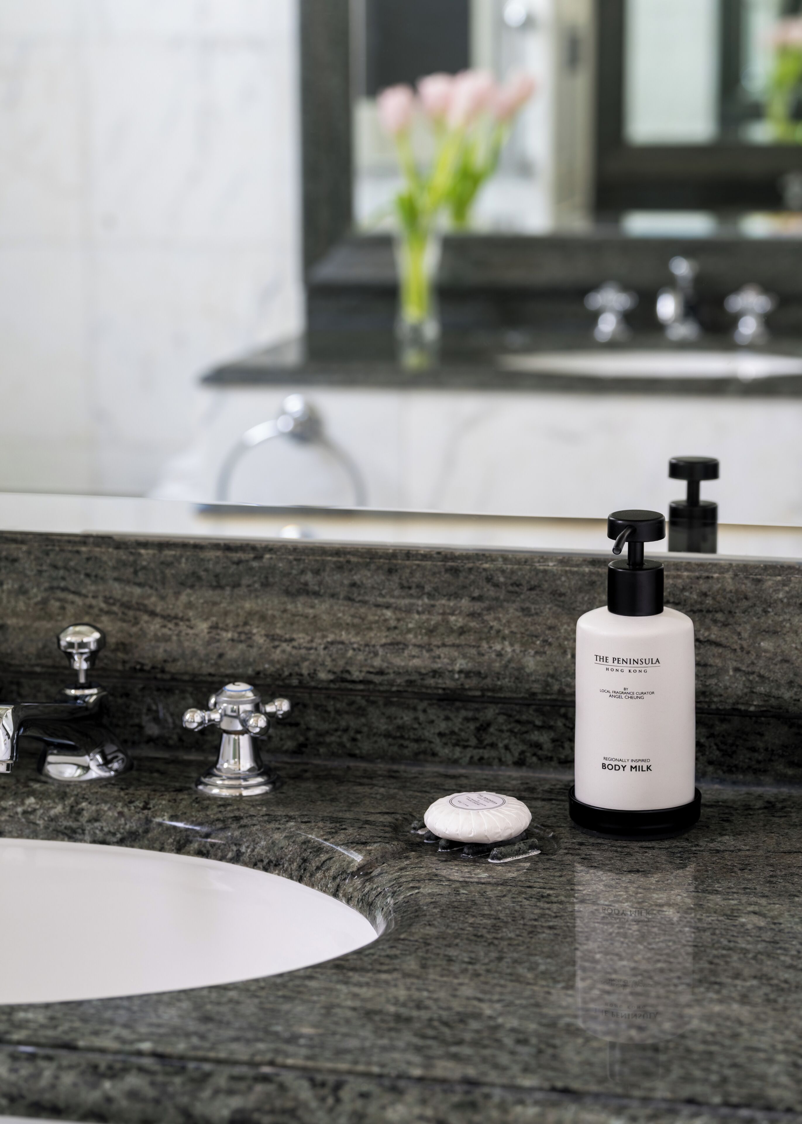 A bathroom with a granite countertop, a white sink, a black bottle of body milk, a soap dish, and a mirror reflecting a vase with pink flowers.
