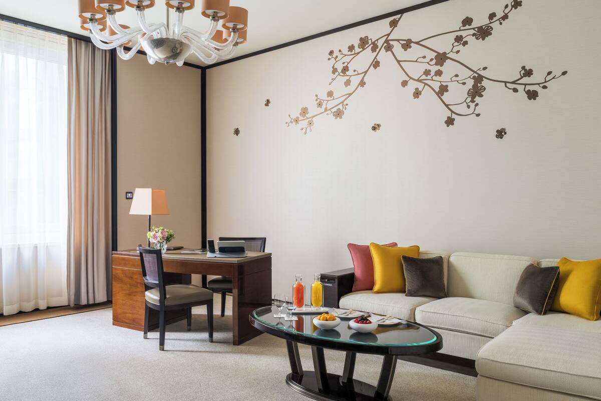 Habitación de hotel con una mesa de comedor, un sofá, una mesa de centro de vidrio y un mural decorativo en la pared.