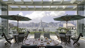 A dining area with a table set for two, two striped chairs, and two umbrellas. A city skyline is visible through large windows. - The Peninsula Hong Kong (Kowloon)