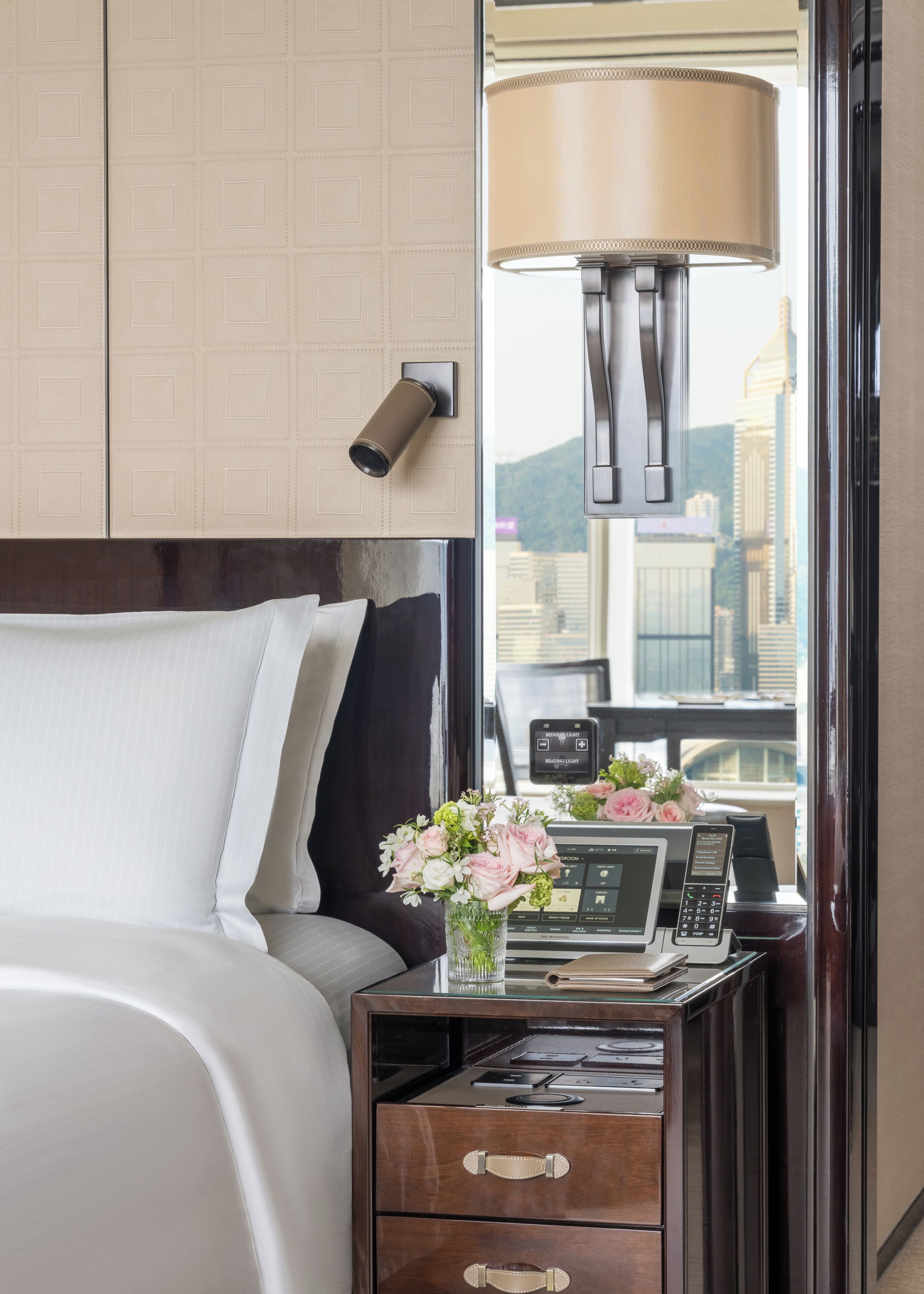 A hotel room with a bed, a nightstand with a lamp, a vase with flowers, a control panel, and a window with a city view.