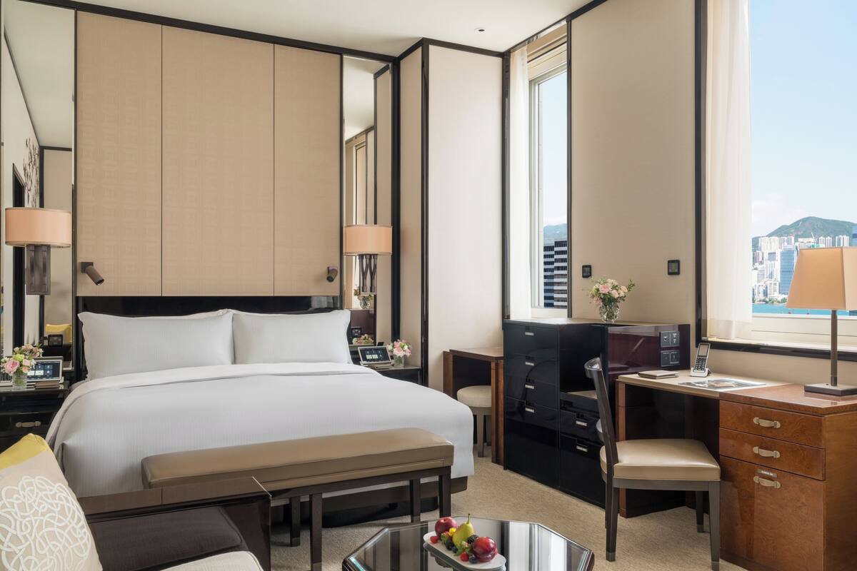 A hotel room with a bed, desk, chair, dresser, and a large window with a city view.