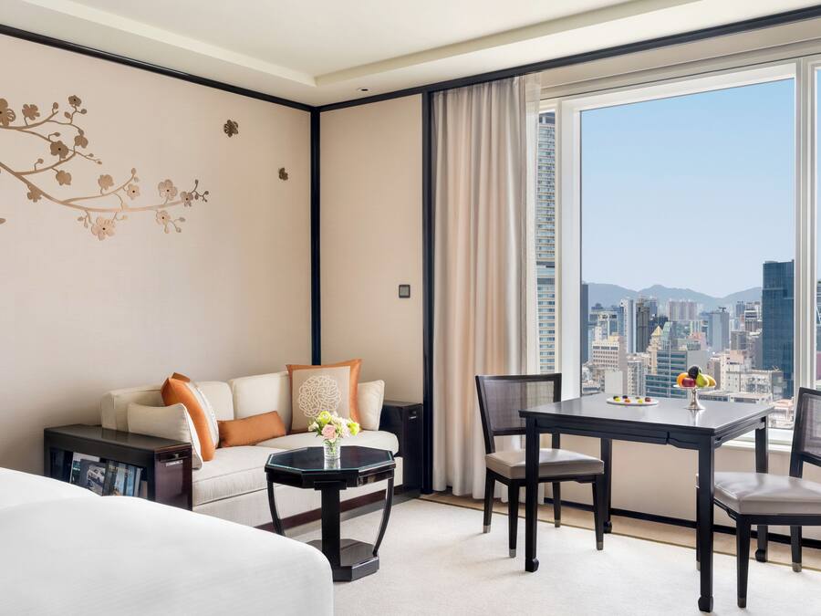 Ein Hotelzimmer mit einem Bett, einem Schlafsofa, einem Esstisch mit Stühlen, einem kleinen Beistelltisch und einem großen Fenster mit Blick auf die Stadt.