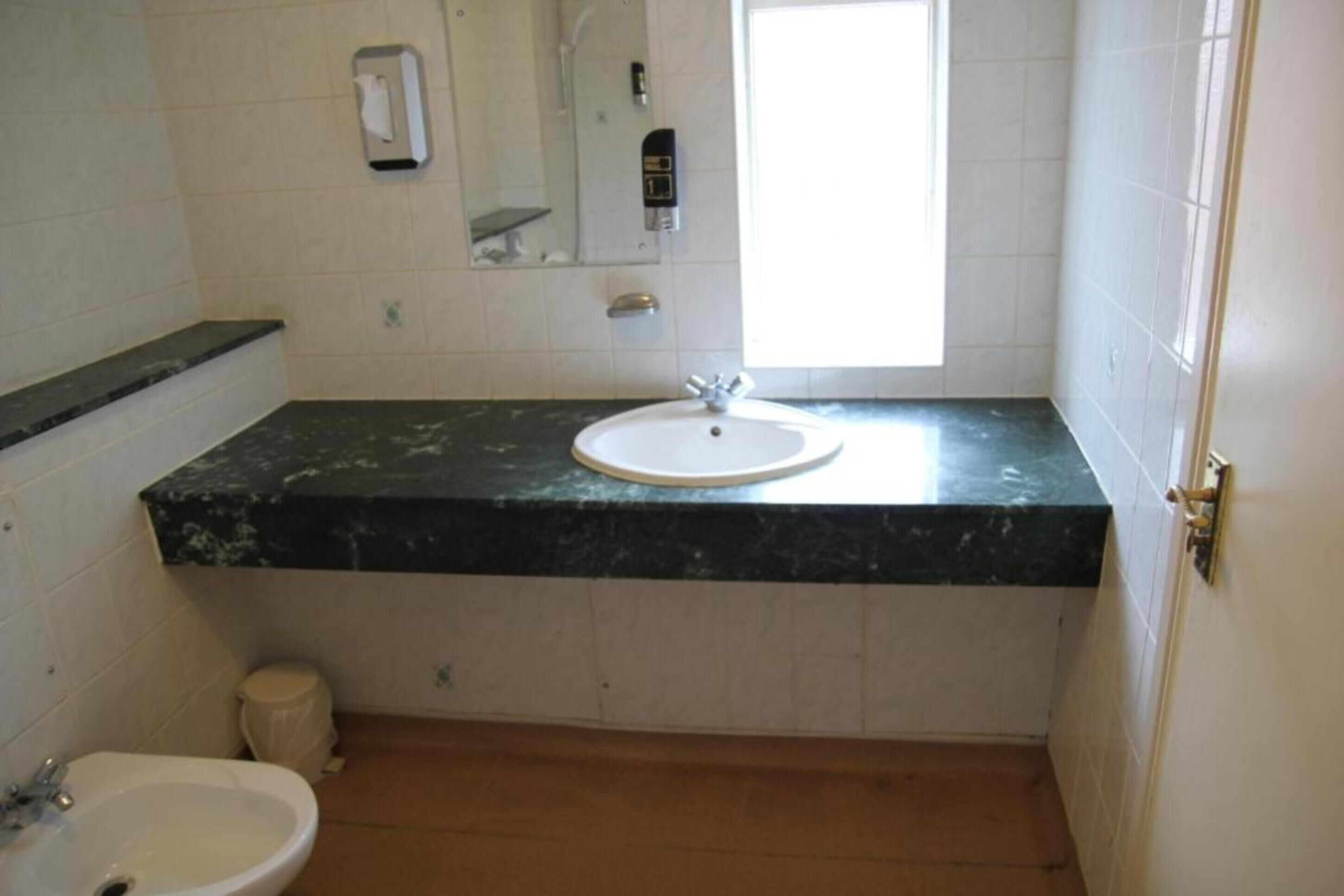 Un baño con lavamanos blanco, espejo, dispensador de toallas y ventana.