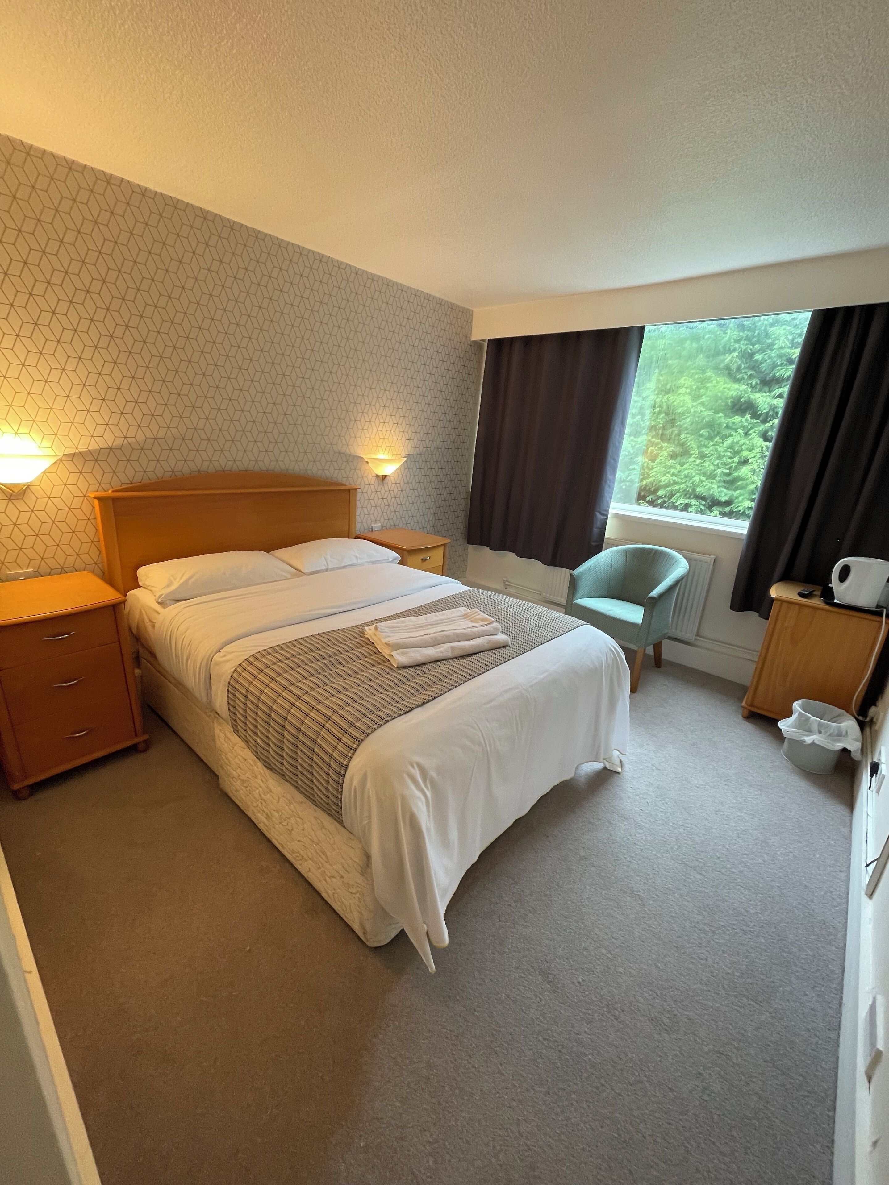 A hotel room with a large bed, two bedside tables, a chair, a radiator, and a window with curtains.