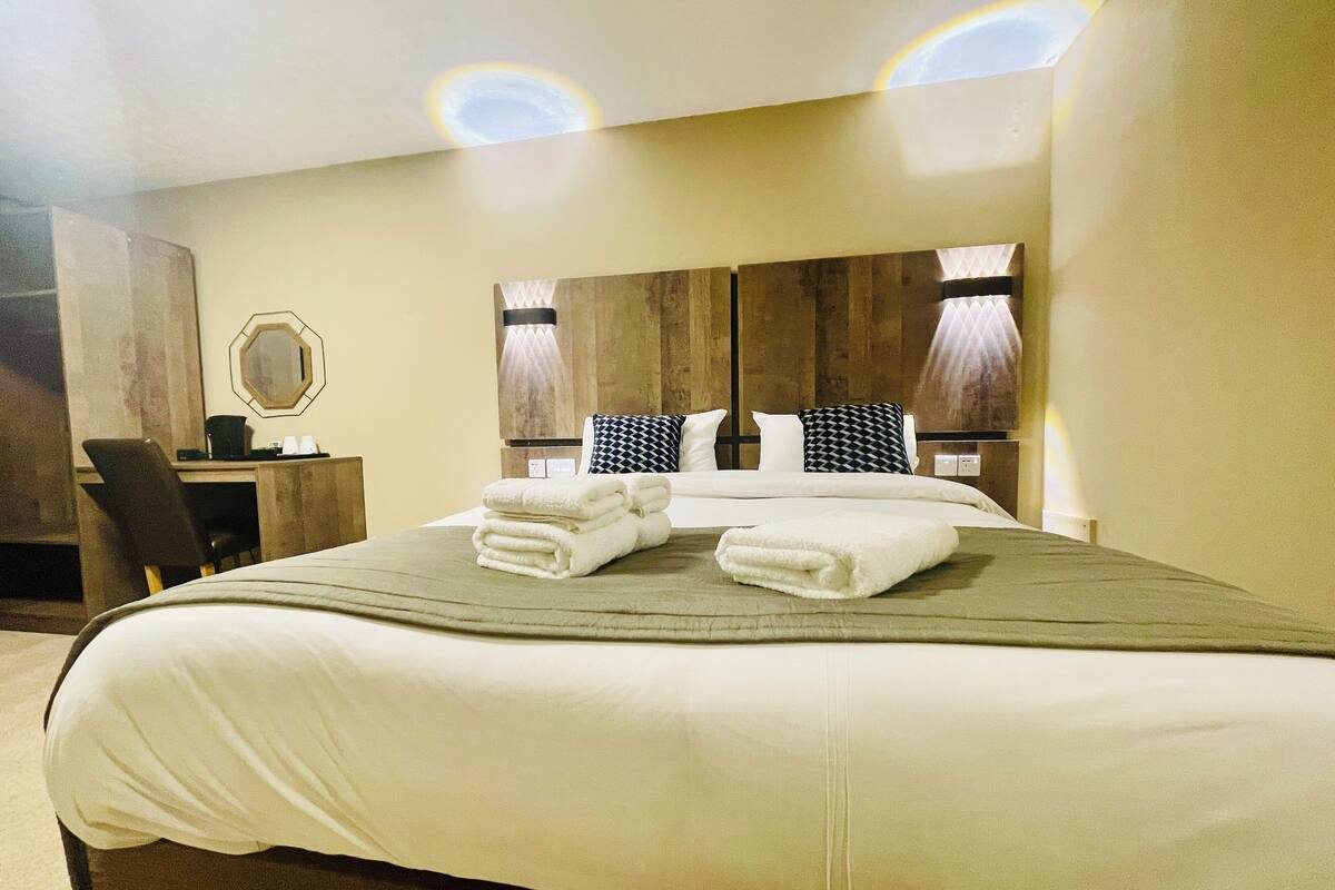 A hotel room with a large bed, a desk, and a chair. There are two lamps on the wall and a mirror above the desk.