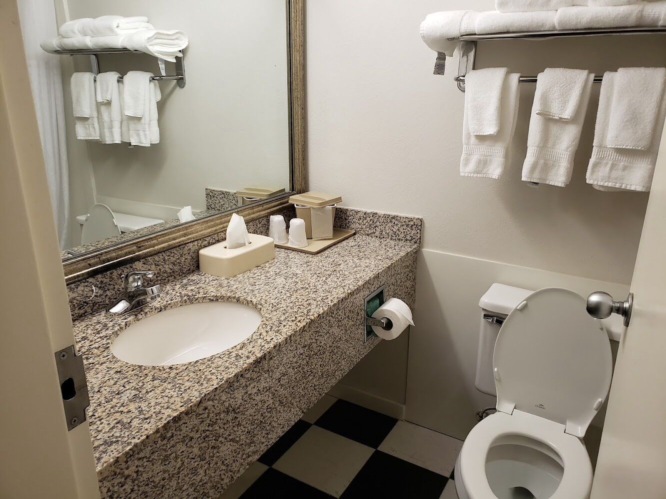 A bathroom with a granite countertop, two sinks, a toilet, and a towel rack with towels.
