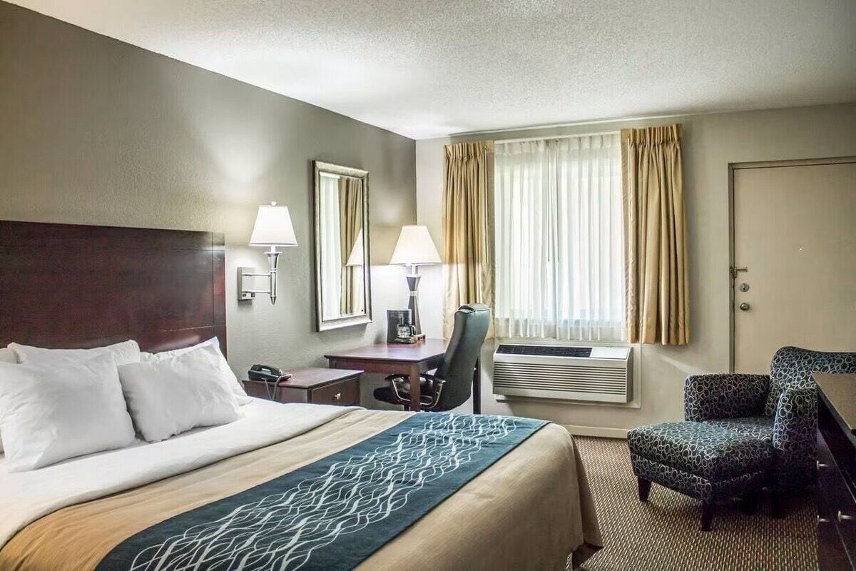 A hotel room with a large bed, two bedside tables with lamps, a desk with a chair, a window with curtains, and an air conditioning unit.