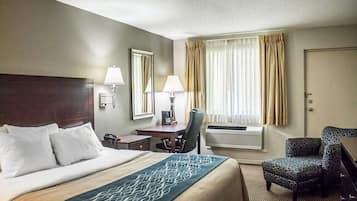 A hotel room with a large bed, two bedside tables with lamps, a desk with a chair, a window with curtains, and an air conditioning unit.