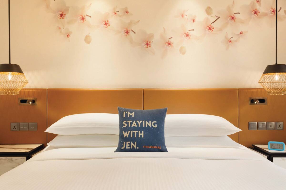 A hotel room with a bed, a decorative wall with floral patterns, two bedside lamps, and a pillow with text.