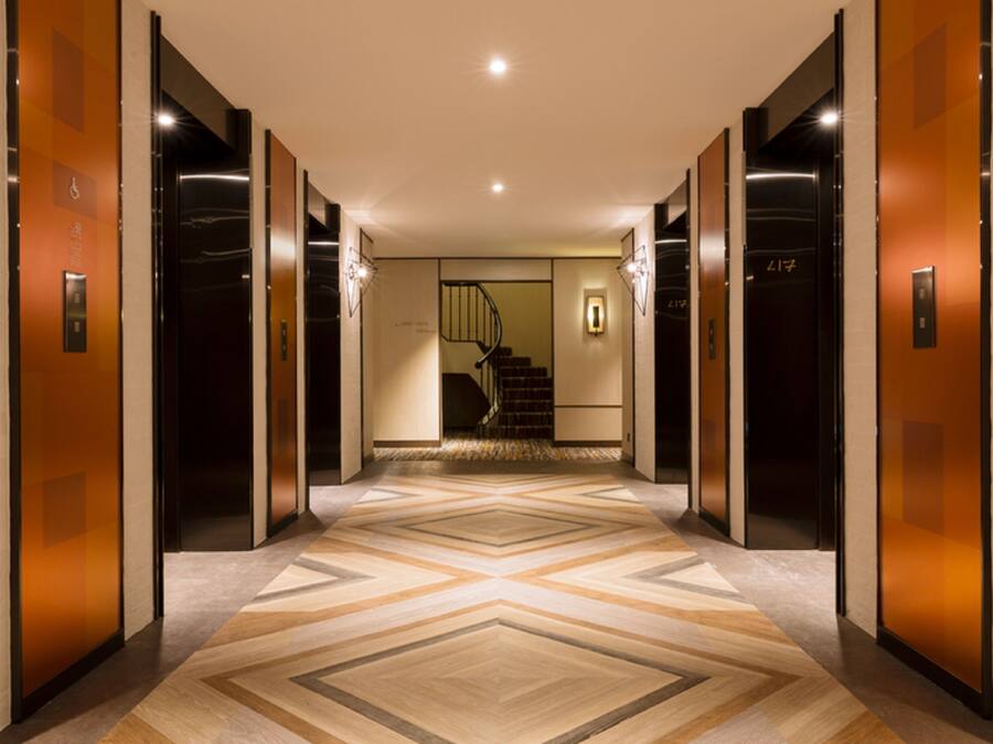 A modern hallway with elevators, a patterned floor, and a staircase in the background.