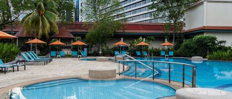 A swimming pool with lounge chairs and umbrellas, surrounded by greenery and modern buildings.