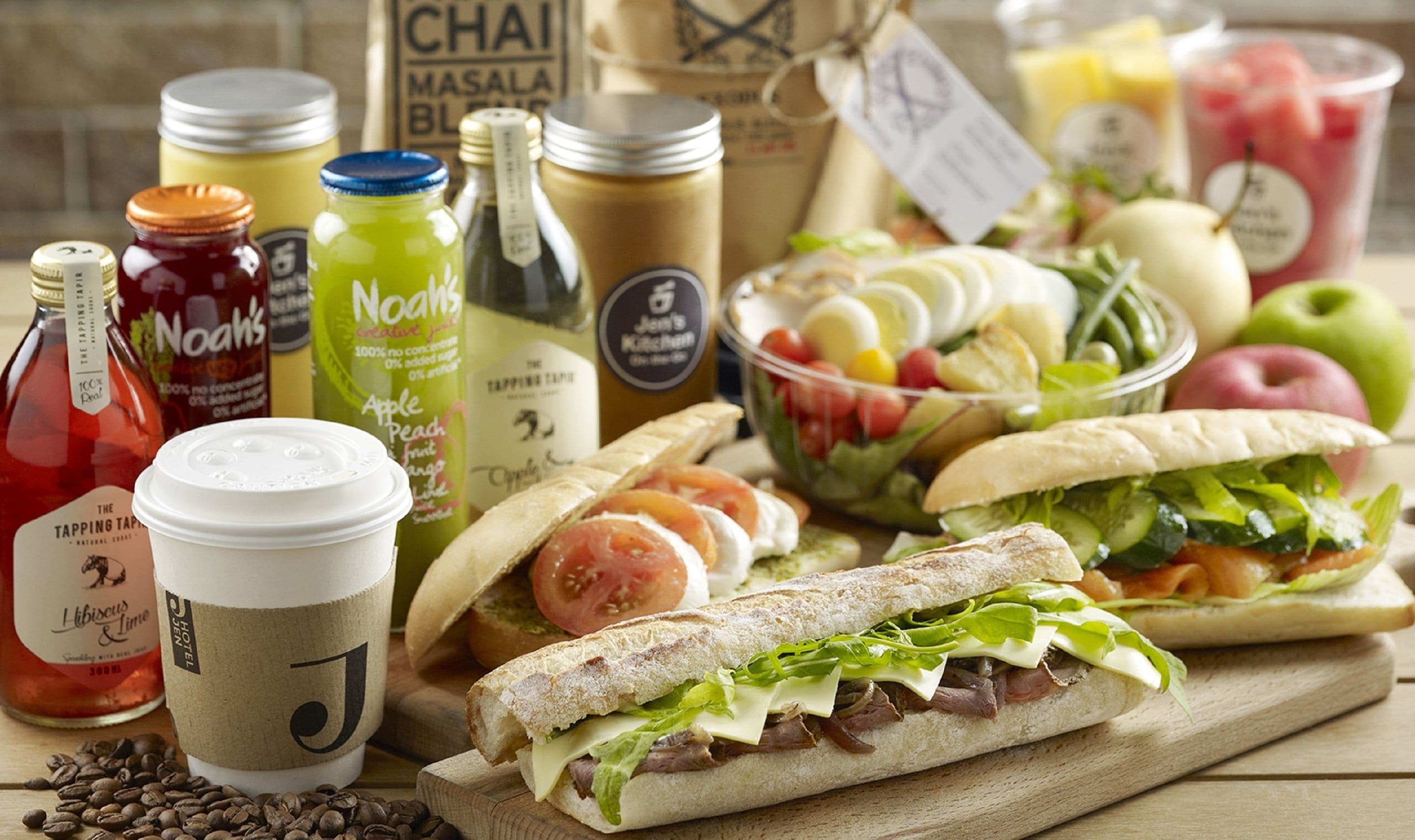 Variedad de alimentos y bebidas sobre una mesa de madera, incluyendo sándwiches, frutas y botellas.