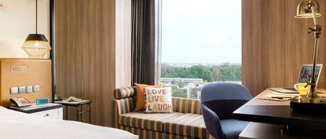Ein Hotelzimmer mit Bett, Schreibtisch mit Lampe, Stuhl, Sofa mit Kissen und Blick ins Freie.