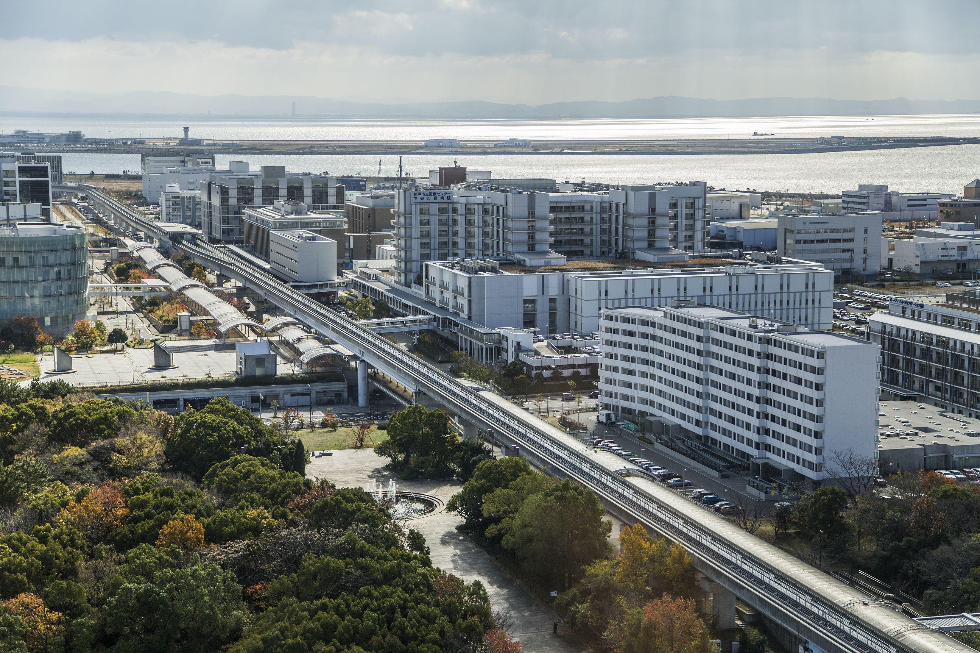 Ein modernes Stadtgebiet mit einer markanten, mehrstöckigen Parkgarage, einem zentralen Fußgängerweg und Blick auf ein Gewässer im Hintergrund.