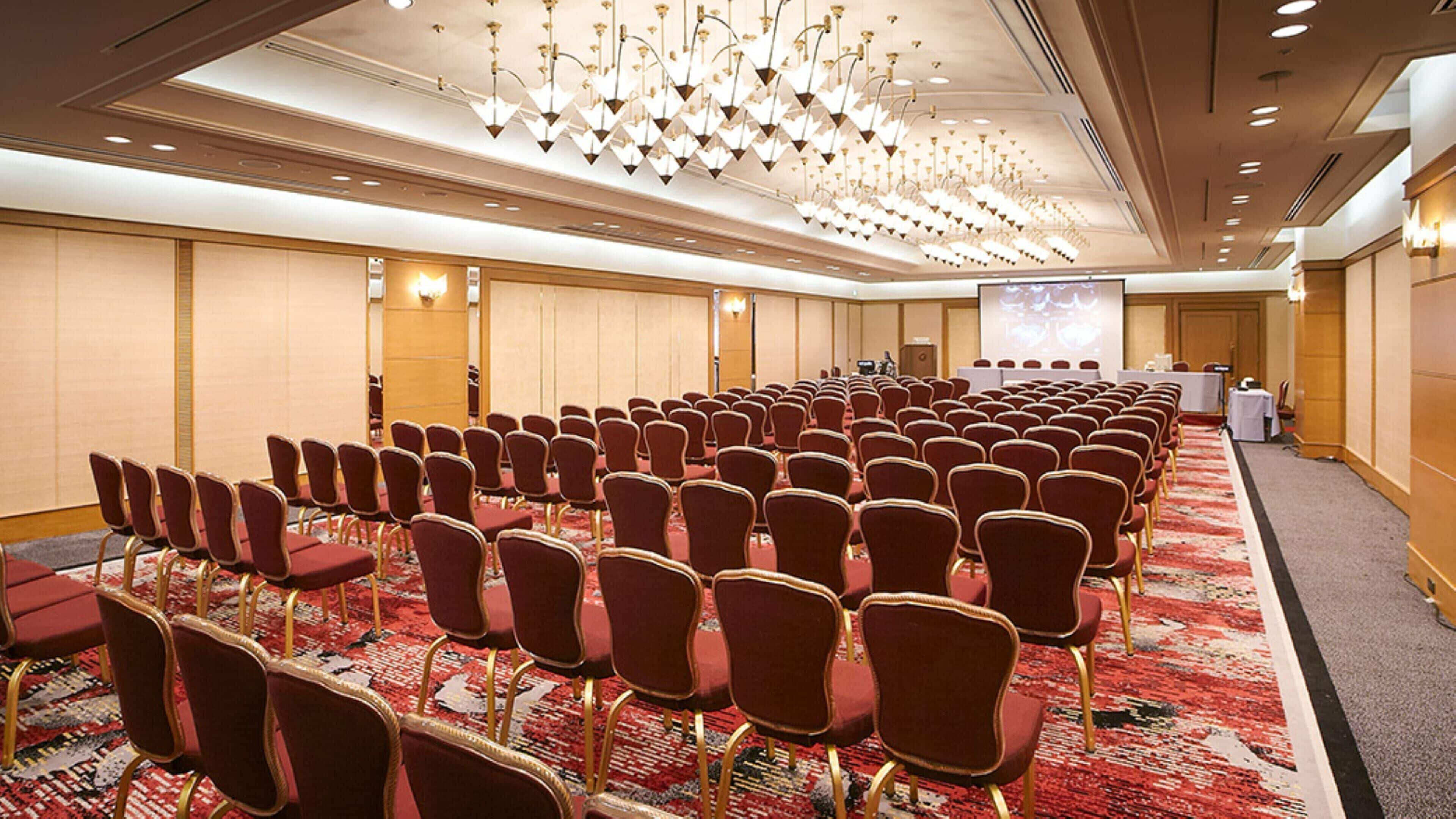 A conference room with rows of red chairs, a carpeted floor, and a large screen at the front.