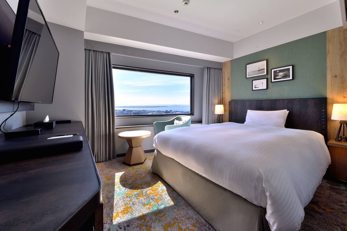A hotel room with a large bed, a desk with a flat-screen TV, a small round table, and a view of the city through a large window.