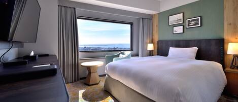 Habitación de hotel con una cama grande, un escritorio con televisor de pantalla plana, una pequeña mesa redonda y vistas de la ciudad a través de un ventanal amplio.