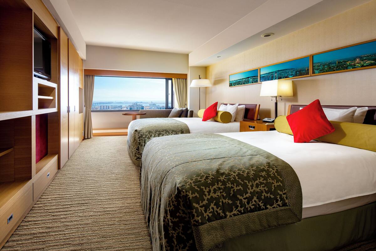 A hotel room with two beds, a large window with a city view, a TV on a wooden cabinet, and a small table with a lamp.