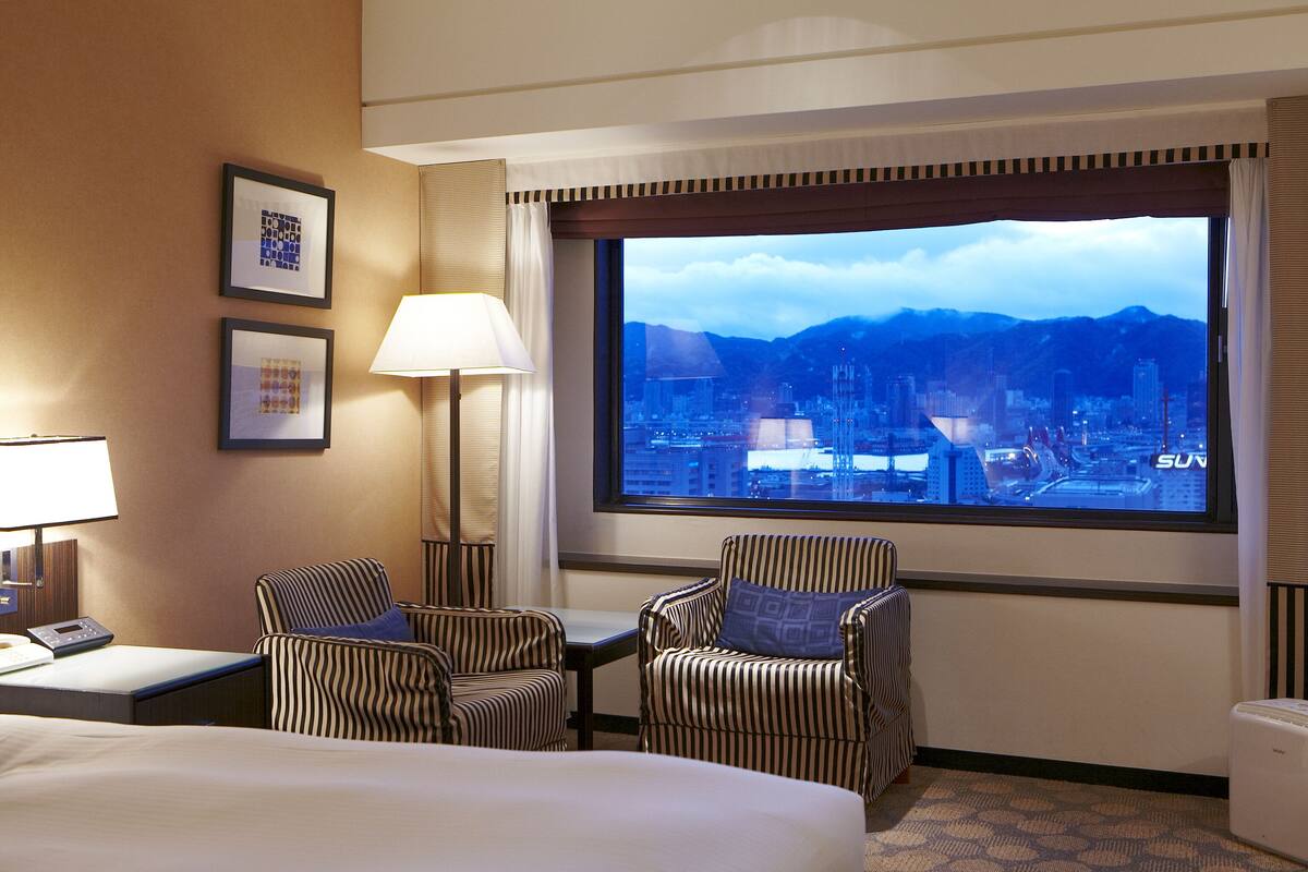 A hotel room with a bed, two striped armchairs, a small table, a lamp, and a large window with a cityscape view.