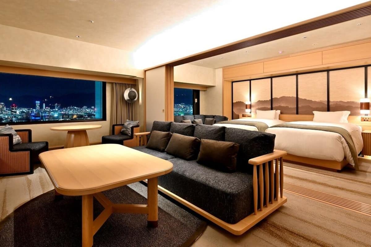 A hotel room with a sofa, a bed, a dining table, and a view of a cityscape at night.