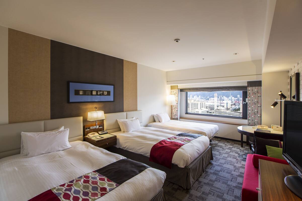 A hotel room with two beds, a desk, a TV, and a large window with a city view.