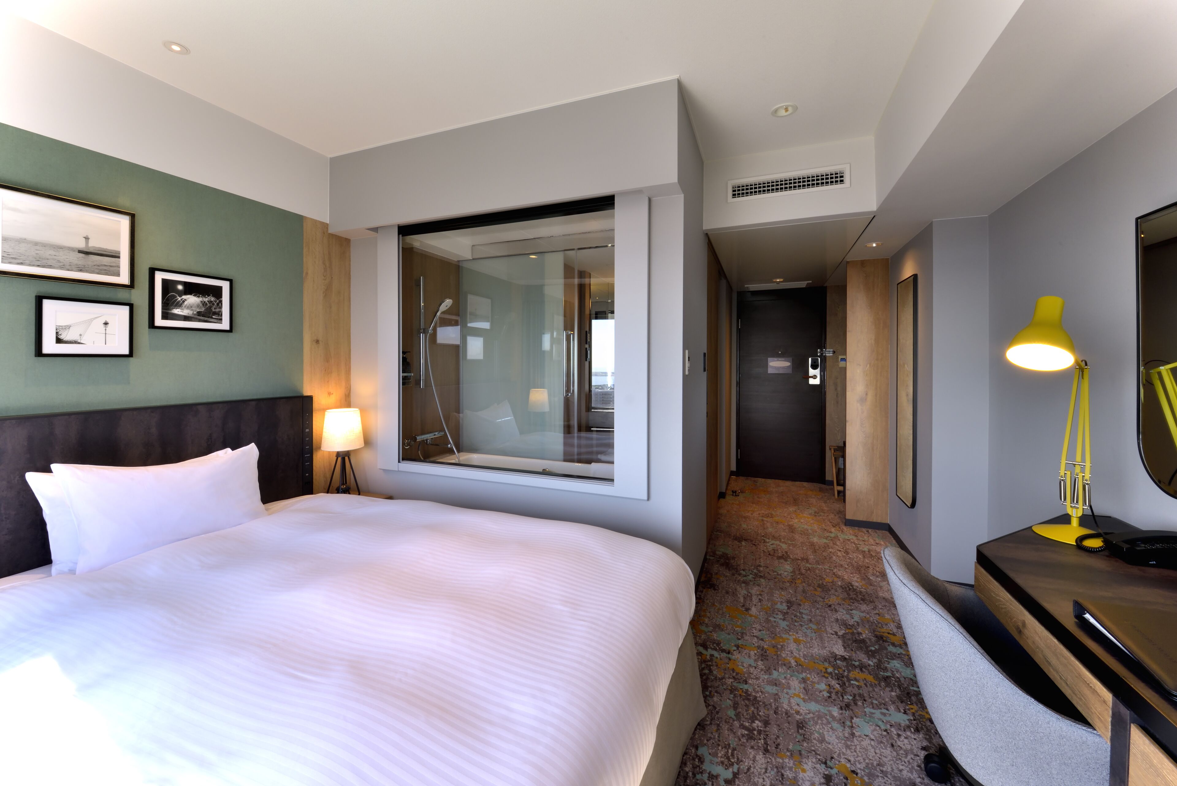 A hotel room with a large bed, a window with a view, a desk with a lamp, and a mirror.