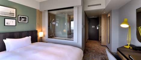 A hotel room with a large bed, a window with a view, a desk with a lamp, and a mirror.